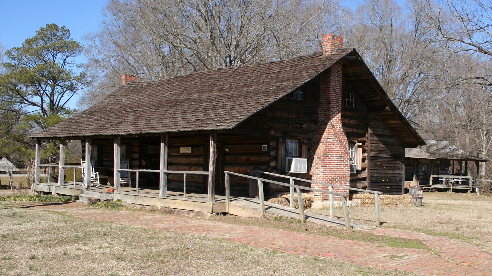 Log cabin with wooden railing surrounding the front porch. The brick chimney is facing the viewer
