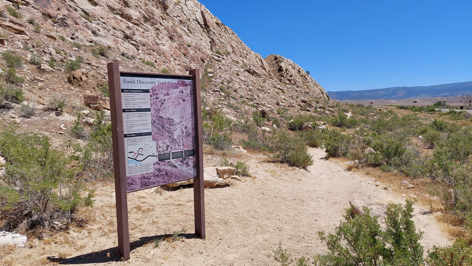A vertical trailhead sign next to a sand trail going along a sandstone cliff face.
