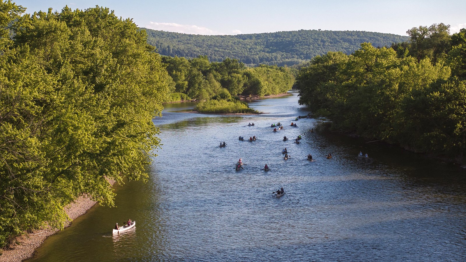A group of canoes on a river flanked by lush green trees and hills under a clear sky.