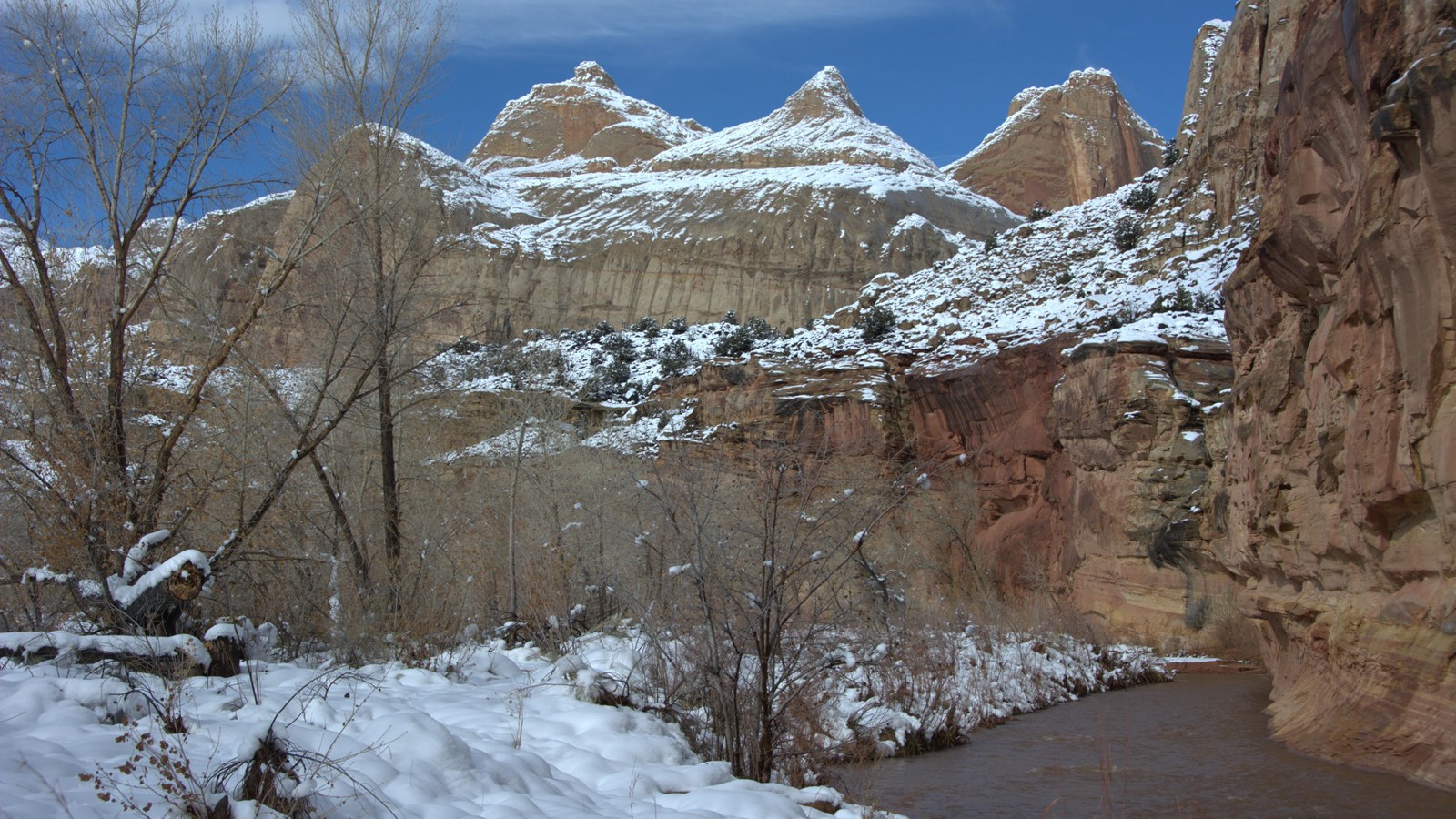 Sandstone peaks rise above a cliff and field of trees along a river, and snow covers the ground