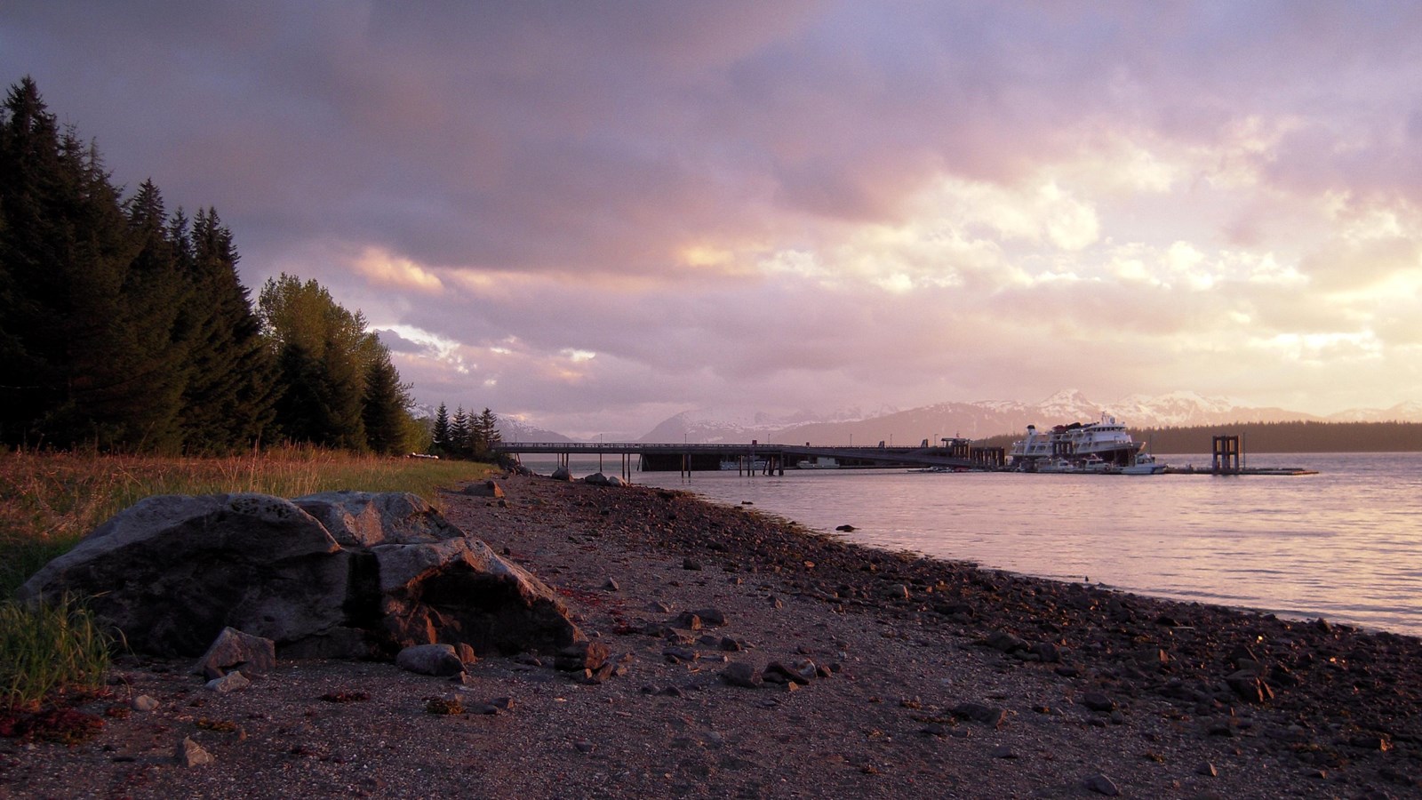 A boulder on a rocky shore, pink sunset clouds overhead. Tall spruce trees flank the shore.