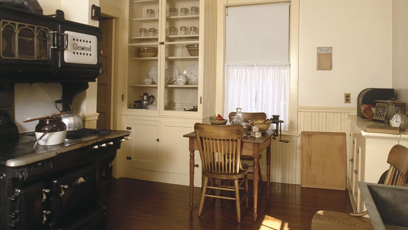 Kitchen with cream-painted cabinets and square work table. Large black stove with kettle and pots.
