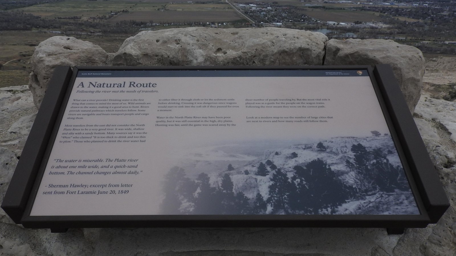 An interpretive wayside sign is anchored to a stone wall overlooking a view of a river.