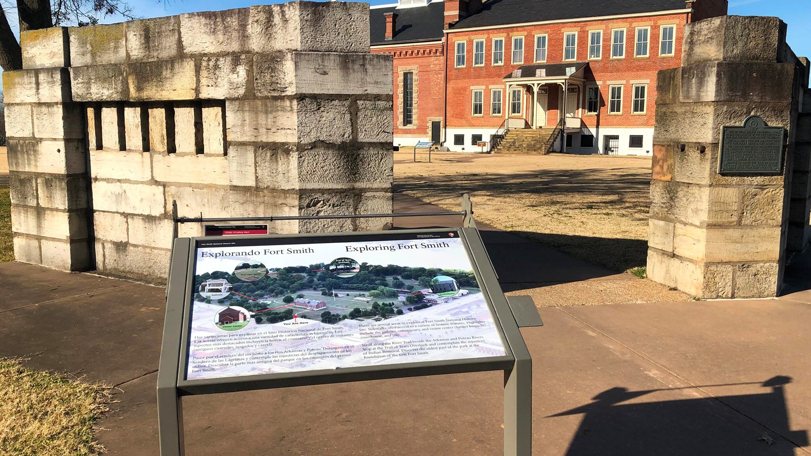 Wayside in front of stone wall entrance with red brick building in background.