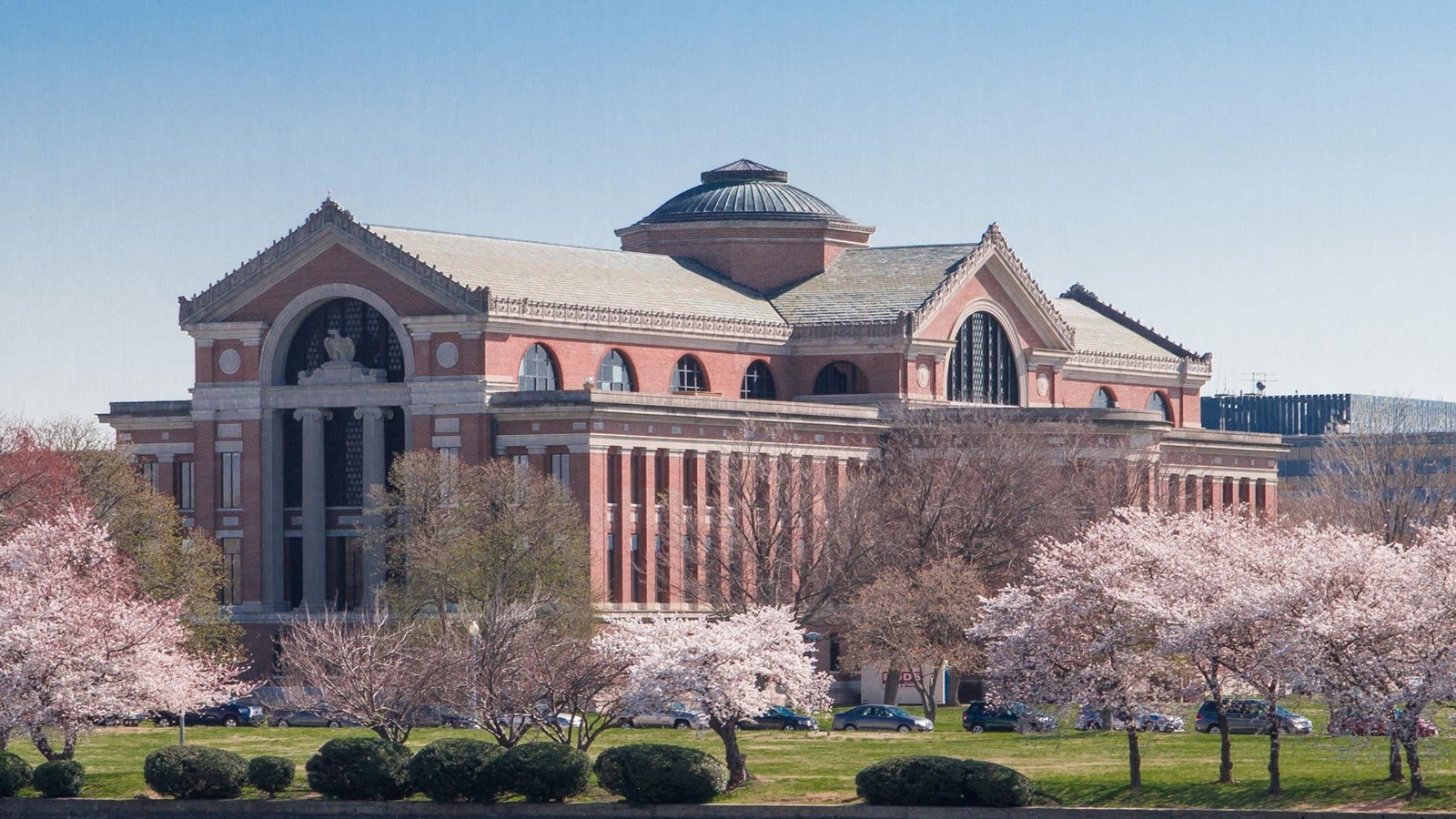 A grand Classical building with archway and central rotunda overlooks a river and cherry blossoms.