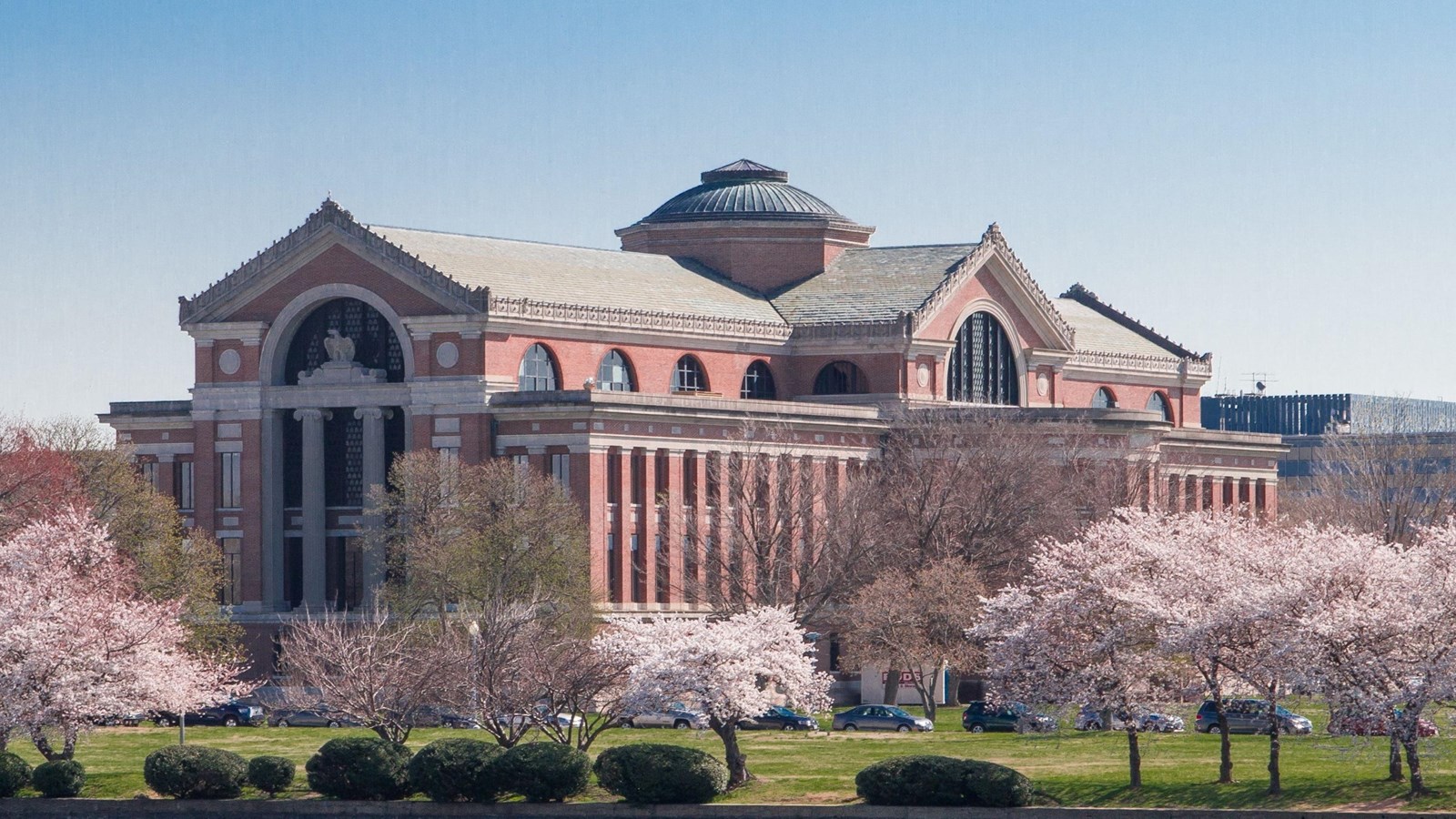A grand Classical building with archway and central rotunda overlooks a river and cherry blossoms.