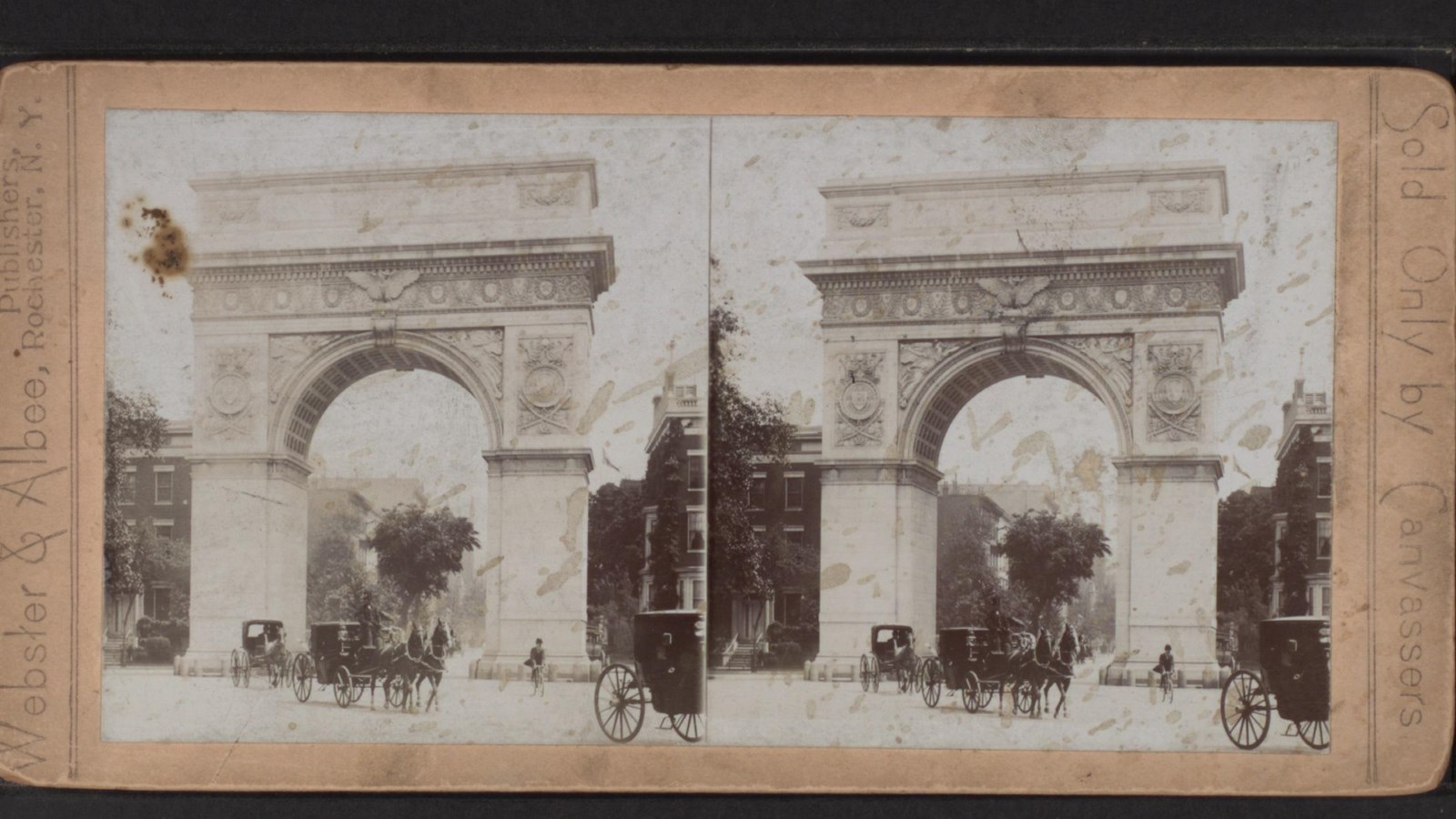 Large decorative arch with horses and buggies passing underneath and buildings in background.