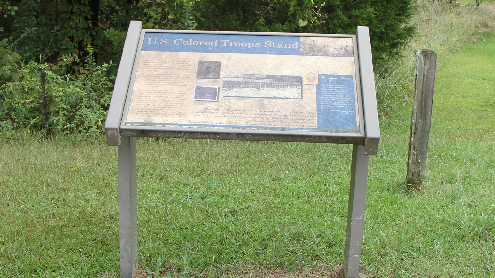 Information panel with brown base behind a paved sidewalk. Mowed grass and trees are behind panel.