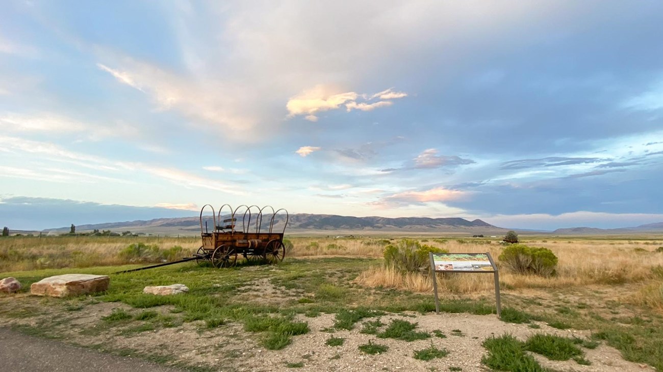 A wayside exhibit sign next to a prairie schooner wagon replica, beautiful sunset clouds.