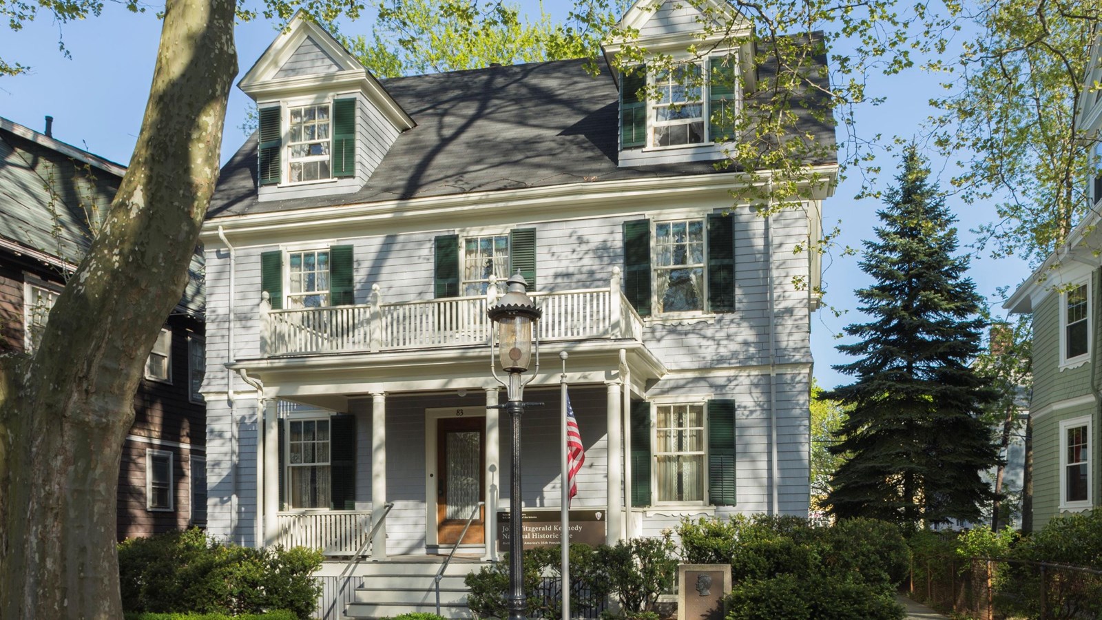 Two story gray house with two dormers in roof. Flag flying on pole in front, gas lamp on sidewalk.