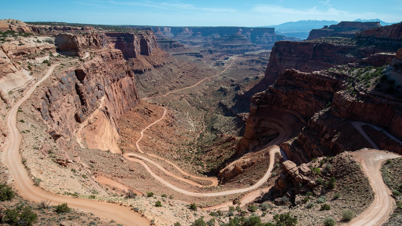 A light beige, unpaved road winds down a steep, multicolored sandstone canyon. 