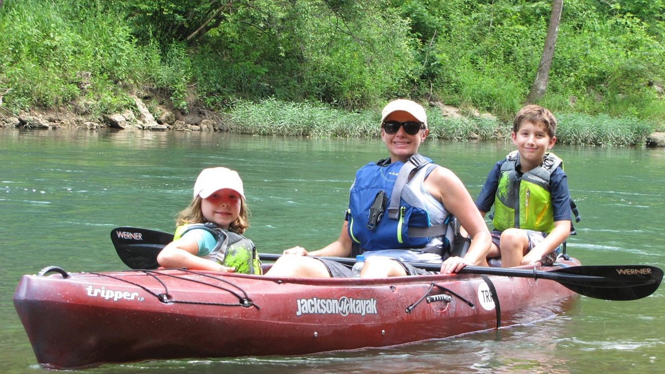 Red kayak on river with woman in middle, girl in front, boy in back, all wearing PFD