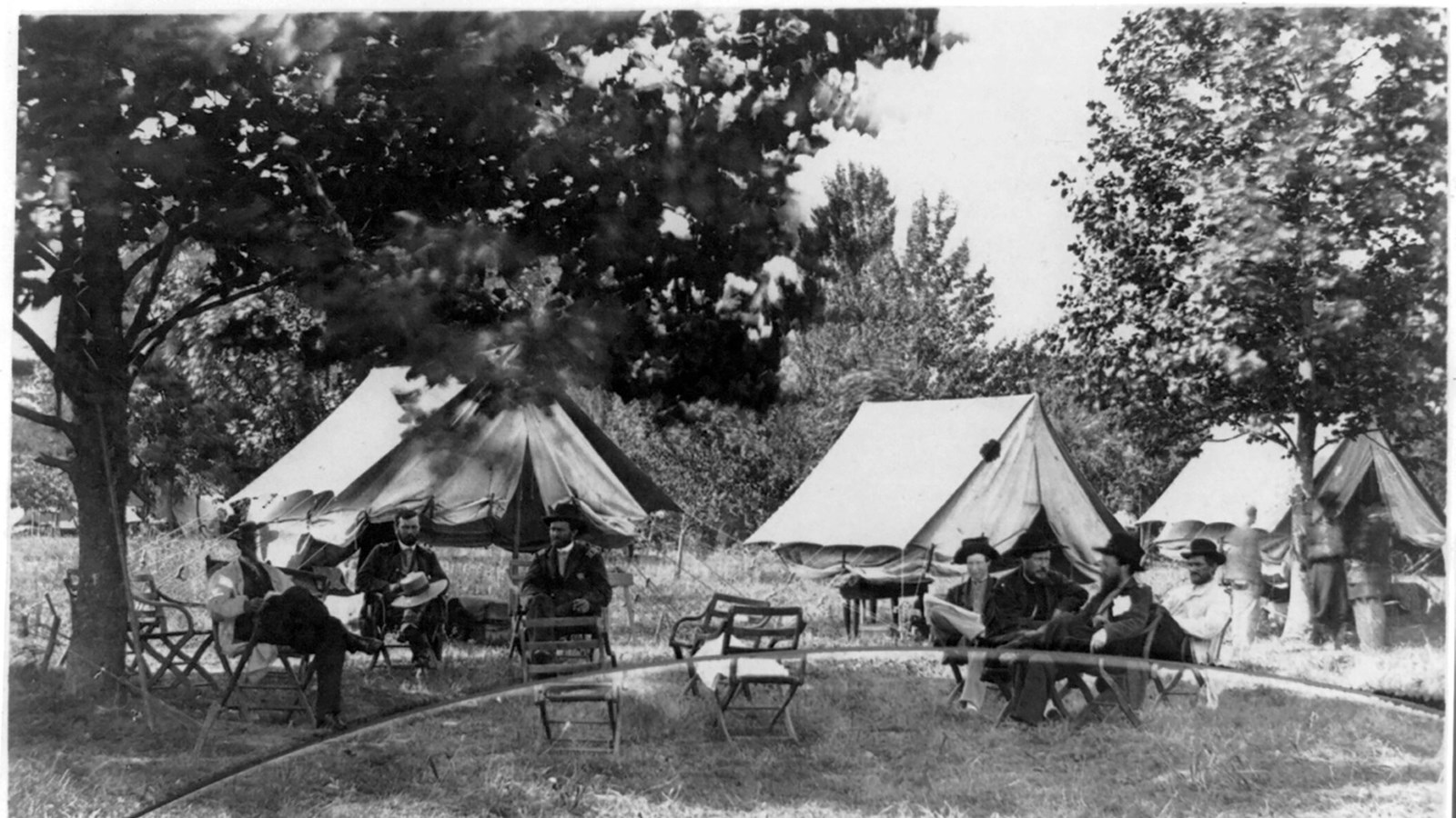 Historic photo from 1864 of men sitting in chairs outside under trees with white tents behind.