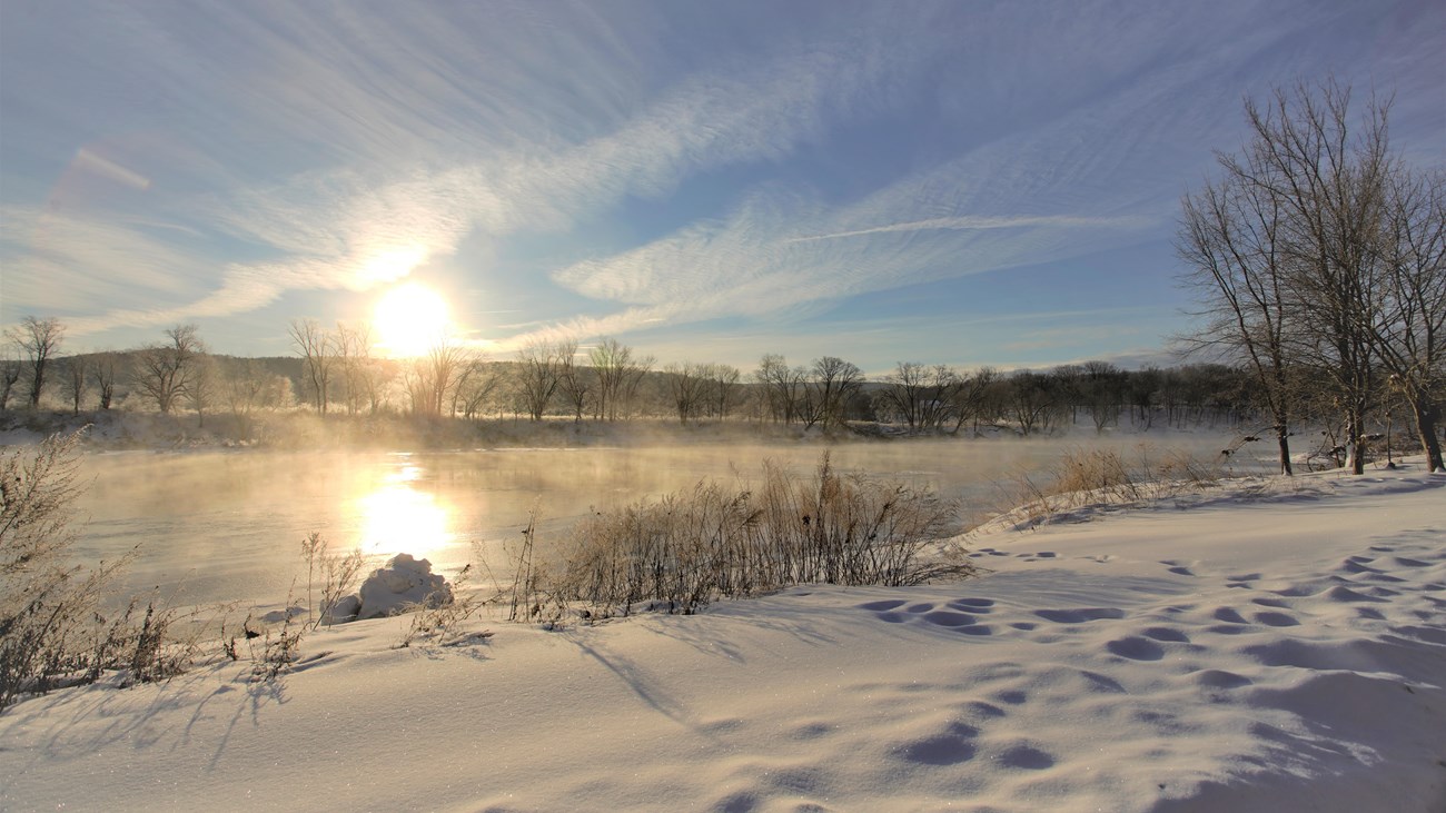 A snow-covered trail looking out at the sun shining on the Delaware River.