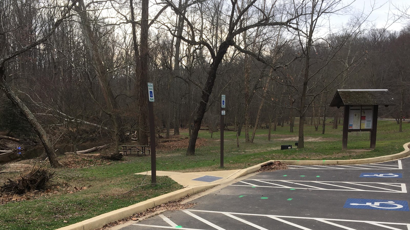 A series of parking spaces and a bulletin board with a table visible by trees beyond it.