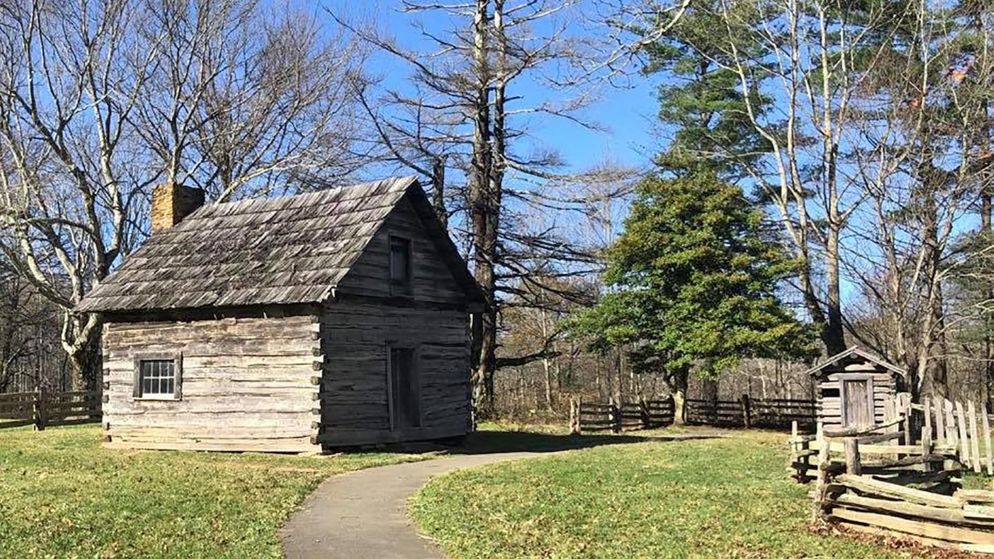 A log cabin and small, log spring house sit on a grassy yard, surrounded by log fences