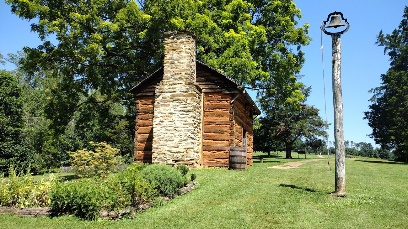 A rustic cabin with a stone chimney and a bell placed on a wooden post.