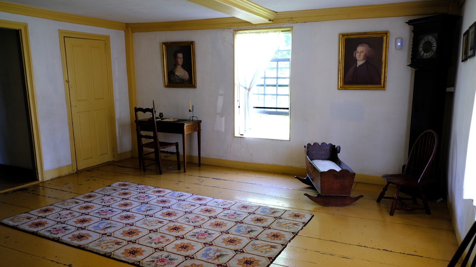 A room with painted floors and walls, a desk, clock, crib, and portraits on the walls.