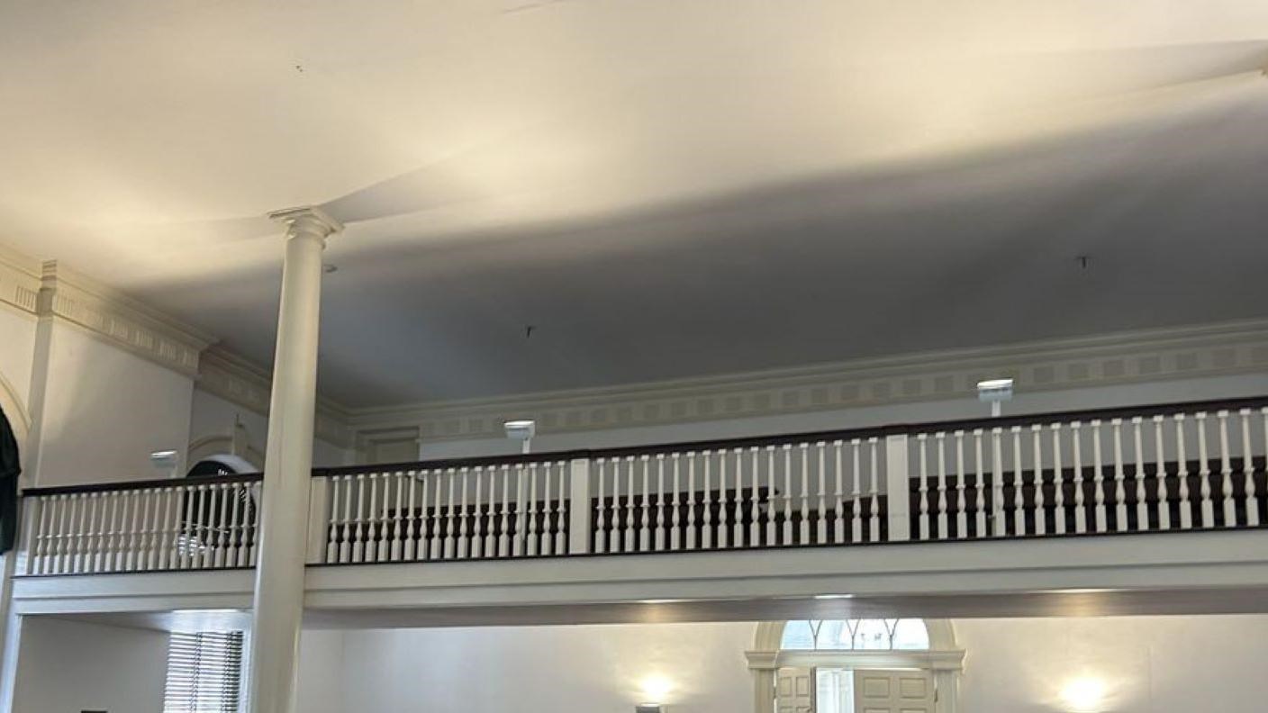 Balcony above a large room, rows of seats with white rails. 