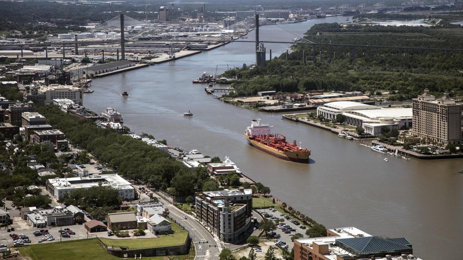 An aerial view of The Savannah River, builings on each side, while a barge moves up river.