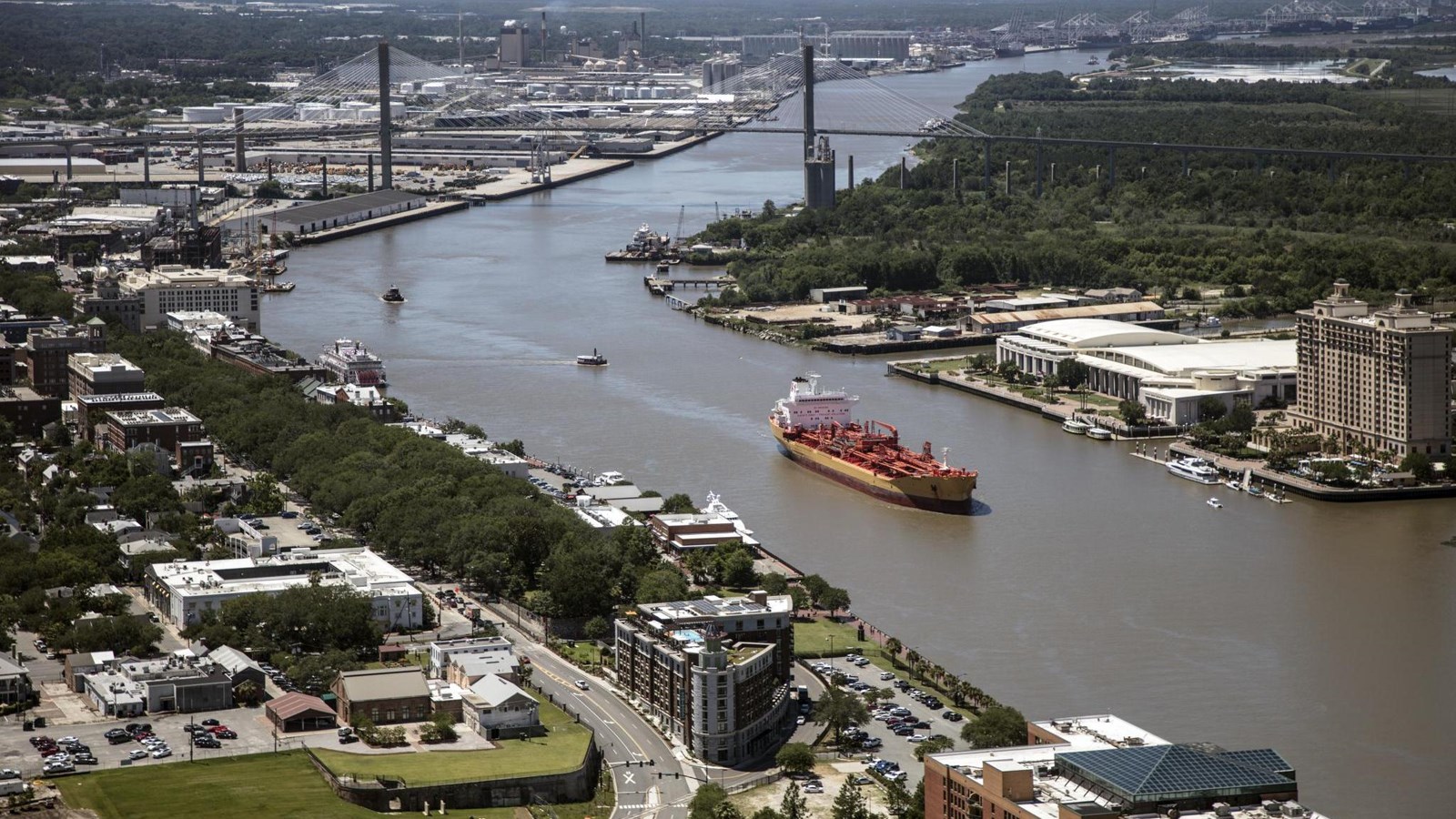 An aerial view of The Savannah River, builings on each side, while a barge moves up river.