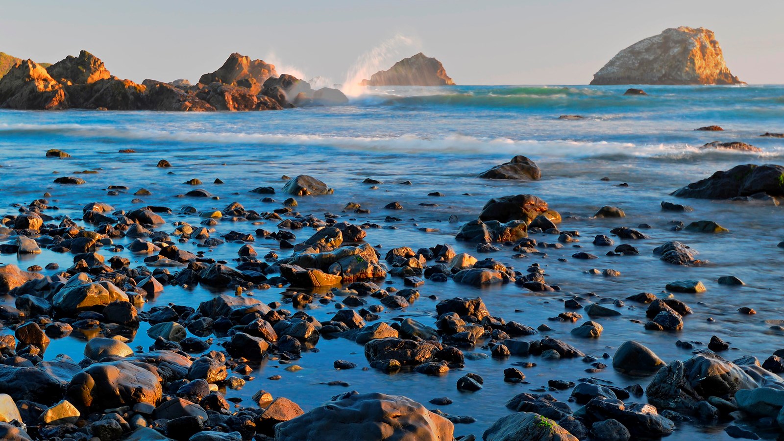 Waves break on rocks illuminated by soft sunset colours.