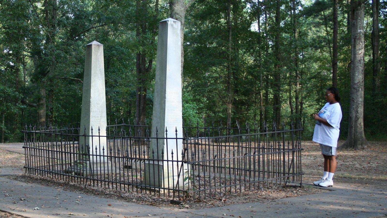 Two white obelisks are surround by iron fence with a park visitor reading the graves