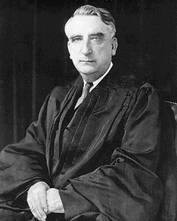 Older white man sits in black robes with his hands clasped in his lap