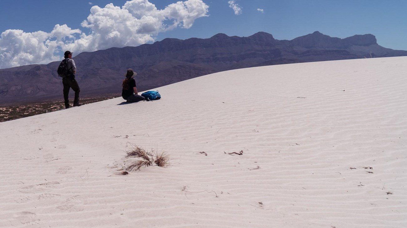 Two people stand on white sand dunes in front of a desert mountain range