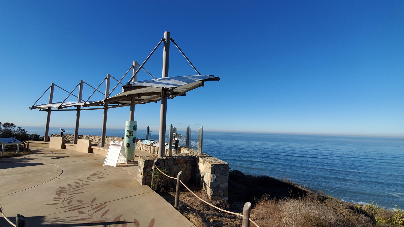 A cement path leads to a lookout covered by white cloth sails. The pacific ocean lies far below. 