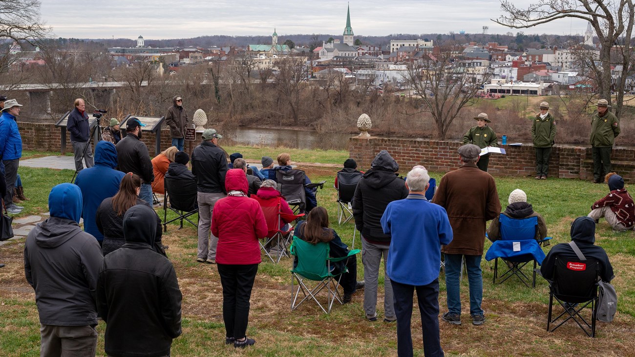 Park rangers speak to a group of people with a river and city in the distance.