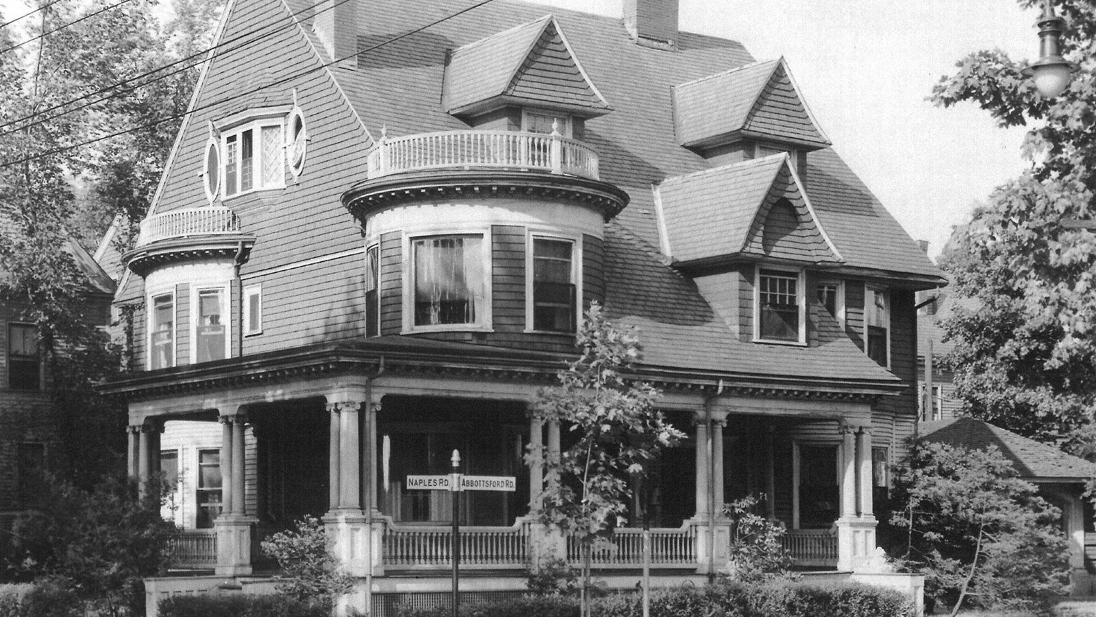 Black and white photograph of three story house with large porch, rounded windows, and dormers