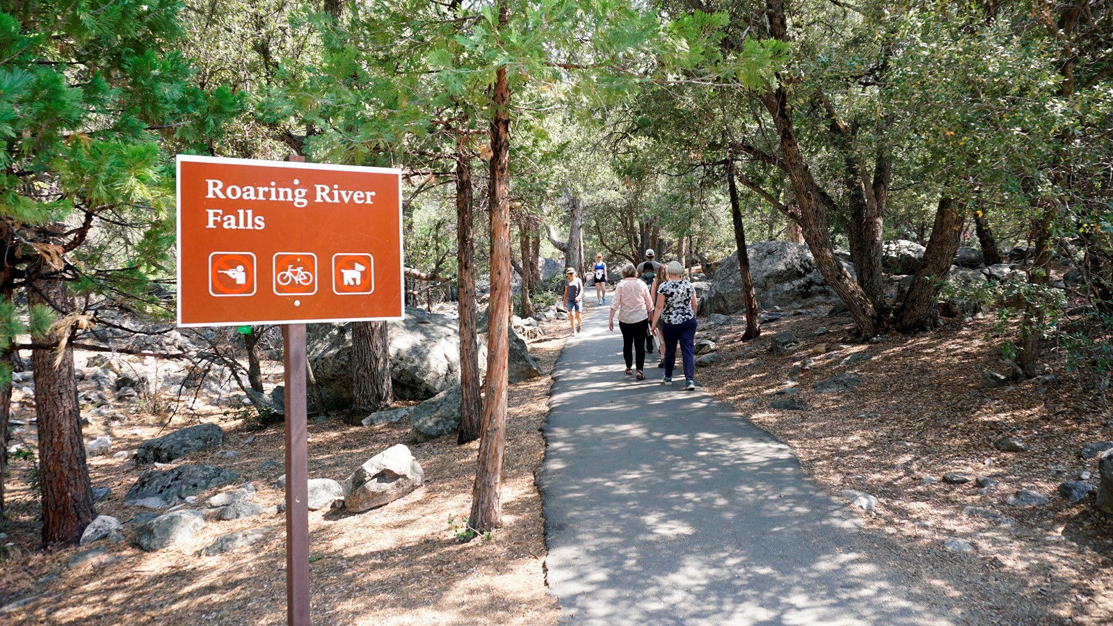 A brown sign with images on it stands next to an asphalt path. The path cuts through trees and rocks
