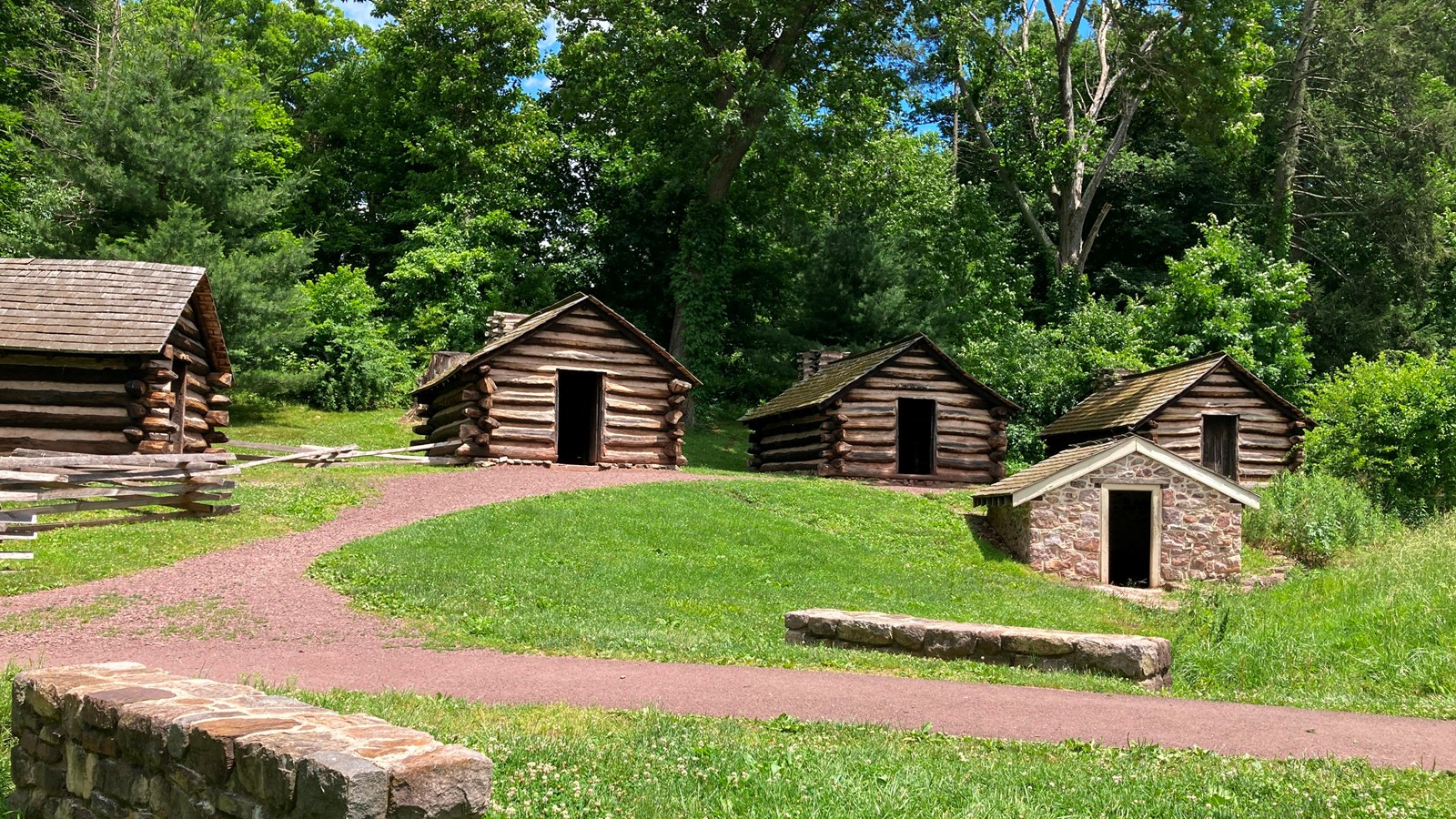 four log huts and a low stone building in front of a dirt path