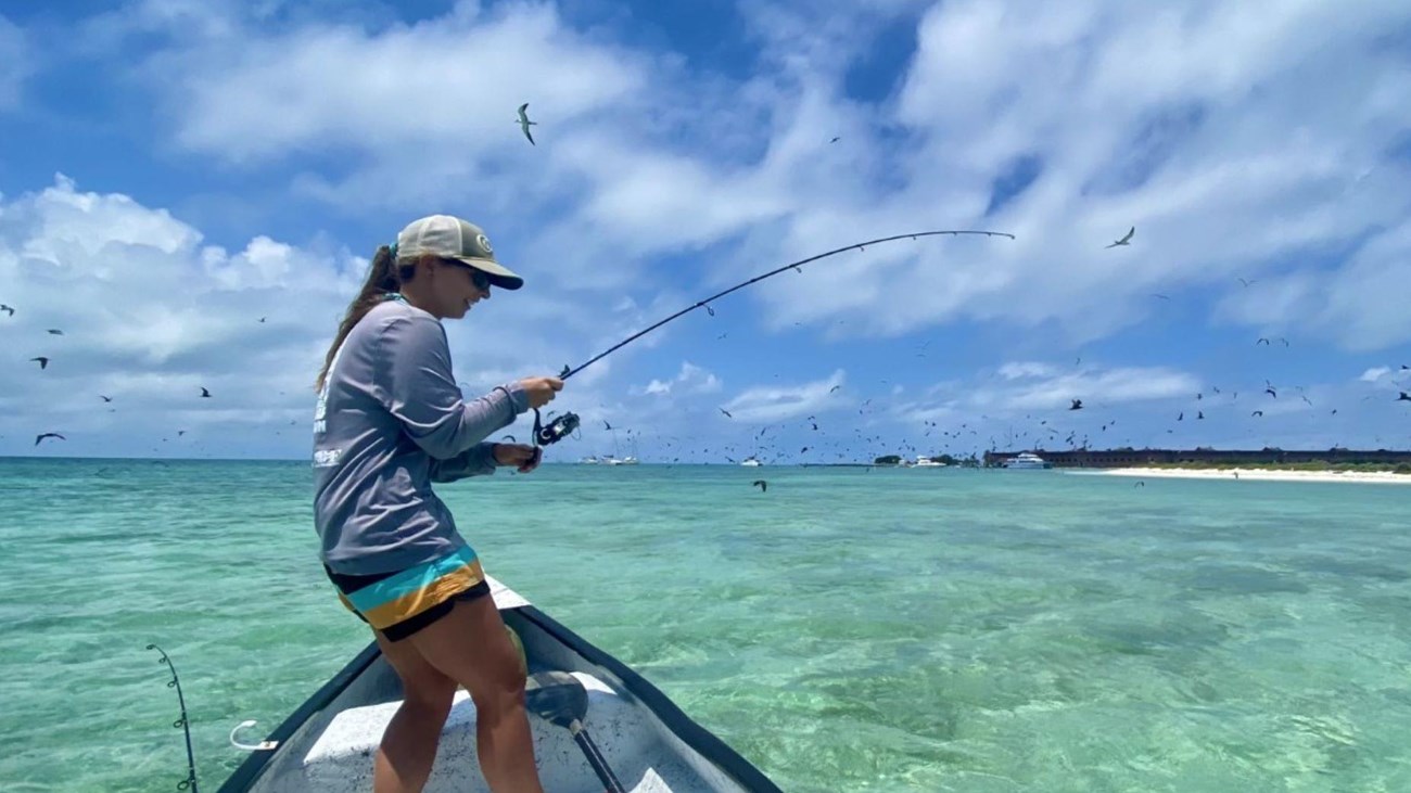 A female angler fights a fish in a small boat atop of crystal clear blue waters.