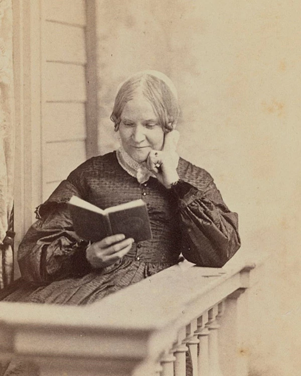 An older woman sits with her hand on her chin, leaning on a balcony while reading a novel