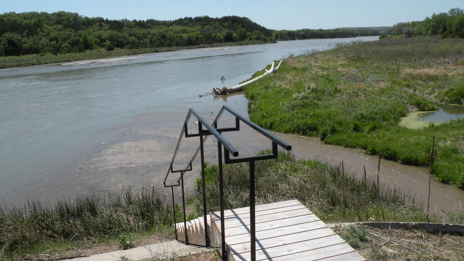 A set of wooden stairs with a railing lead down to a riverside and landing.