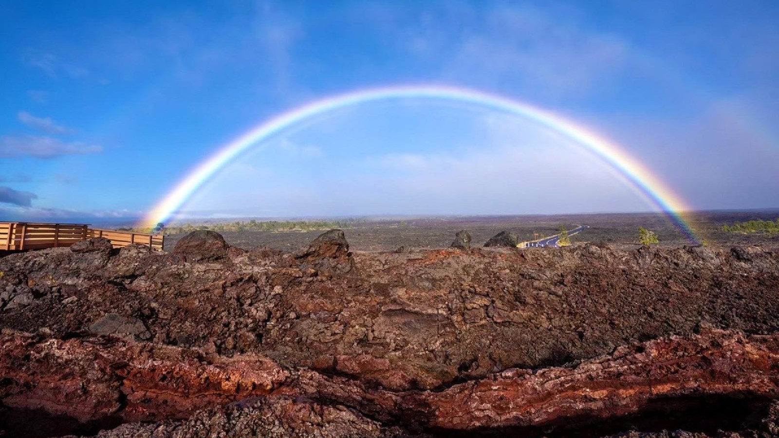 A wooden overlook in a volcanic lava field with a rainbow in a distance. 