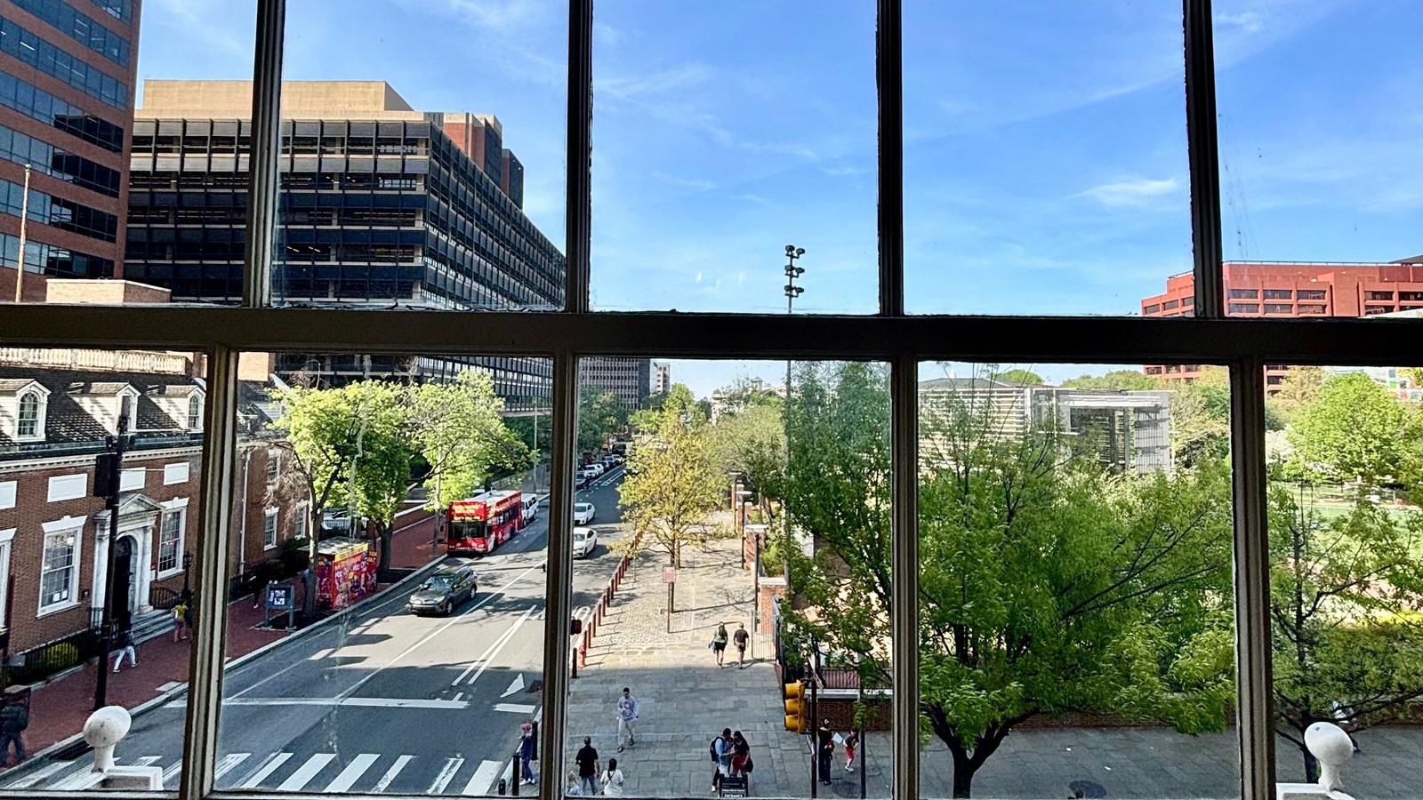 Looking through a large window at a city street. Trees, people, cars are on the street.