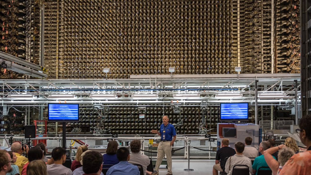 A group of people sit in chairs as a man gives a talk in front of a large industrial apparatus.