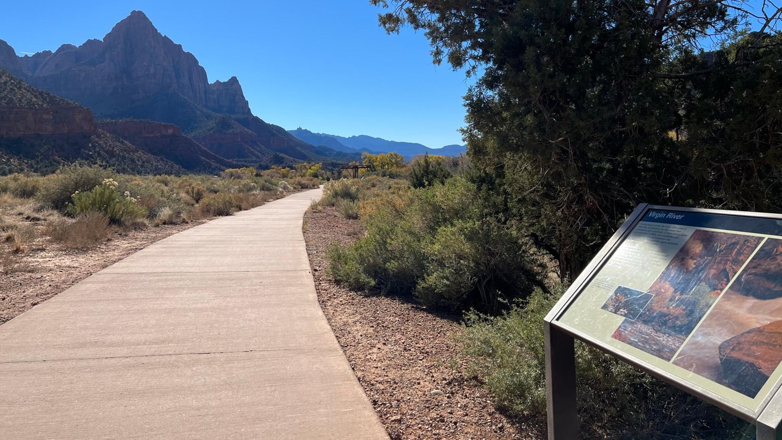 Photo of a sign to the right of a paved trail. A triangular mountain is visible in the background. 