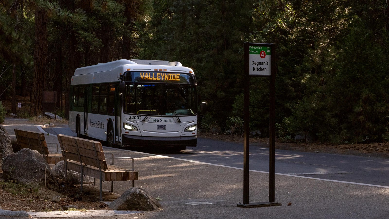 A white bus approaches a sidewalk with benches nearby