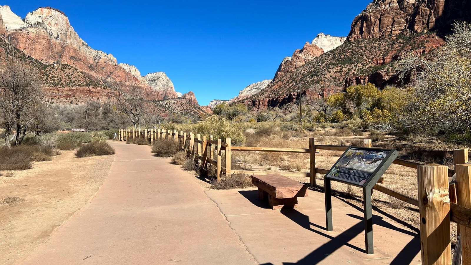 A split-rail wooden fence running along a paved trail. Mountains are visible in the distance