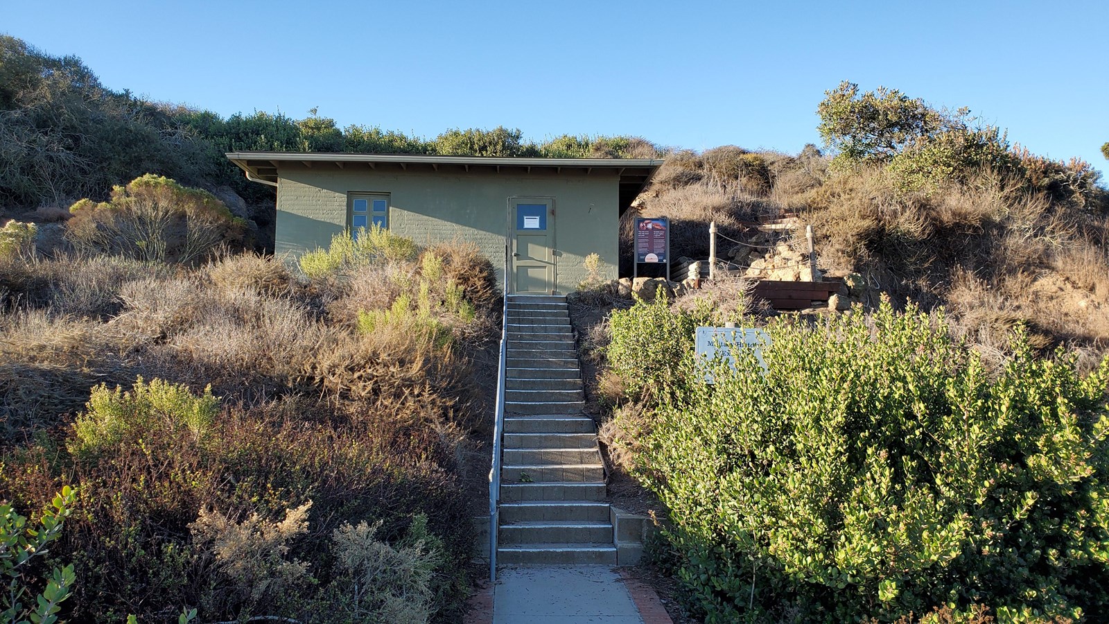 A gray staircase leads up to a rectangular olive green building embedded in a brownish hillside.