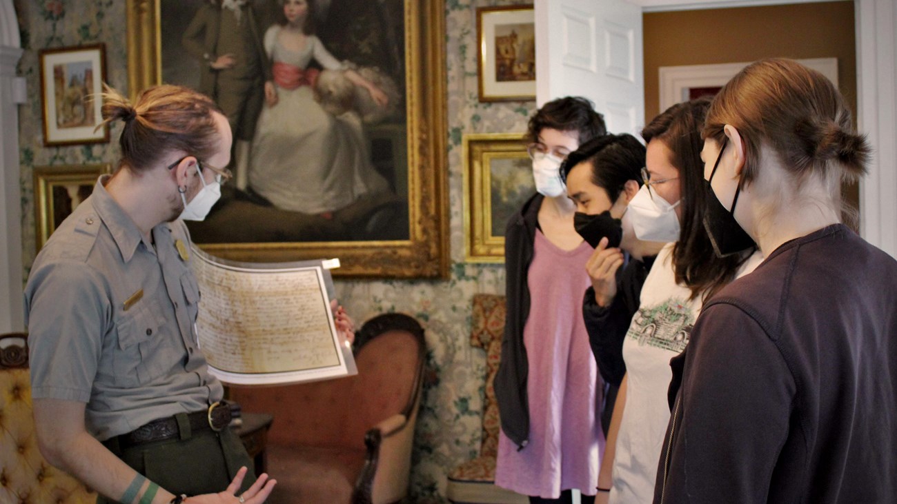Ranger showing four visitors a map in a Victorian parlor with large oil painting in background