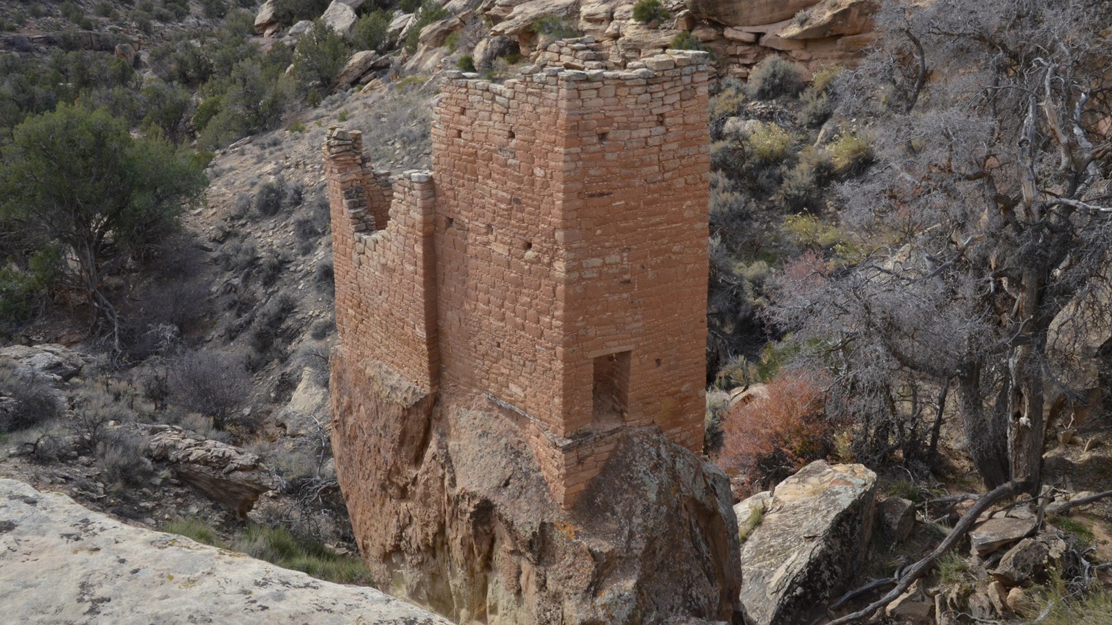 a tall, rectangular stone structure perched on a boulder in the center of a shallow canyon.