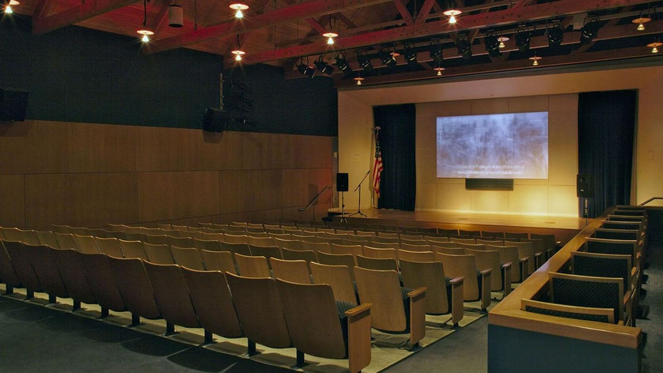 An auditorium with wood wall, exposed truss ceiling, and stage with screen.