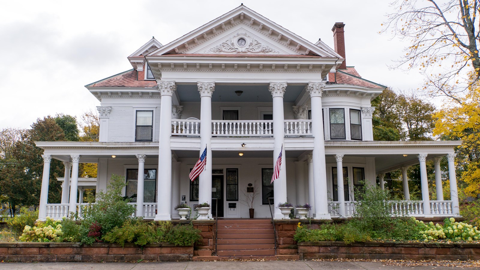 Fall scene of large three-story white house with grand entrance, wrapping porch, and brick chimney.