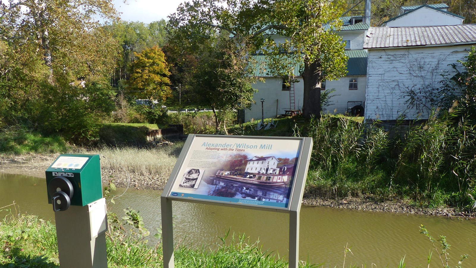 A graphic panel and a hand-cranked device stand beside a waterway with white buildings and a road on