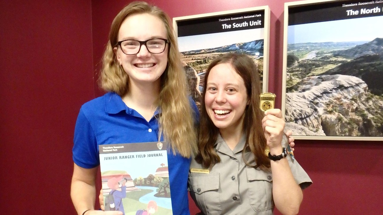 Two women smiling at the camera, one in a ranger uniform, holding a junior ranger book and badge.